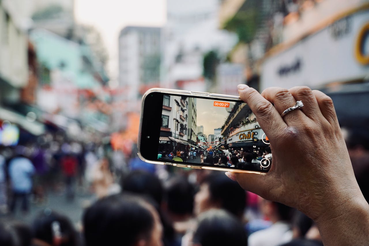 Hand holding smartphone capturing bustling city street scene with crowd.